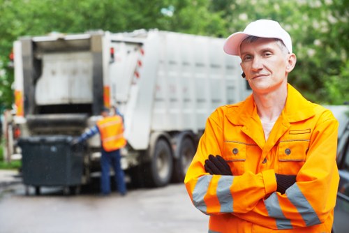 A professional waste management worker wearing a bright orange high-visibility jacket with reflective stripes and a white cap stands with arms crossed in front of a large, grey, industrial rubbish collection vehicle parked outdoors on a driveway or paved area. In the background, another worker in similar high-visibility clothing is seen operating the rear loading compartment of the truck, which has its rear doors open, revealing black waste bins inside. Surrounding the scene are lush green trees and foliage, suggesting a suburban or urban environment. The overall lighting indicates daytime with natural light illuminating the scene, emphasizing the textures of the technical equipment and the outdoor setting. The image portrays a waste collection or rubbish removal operation, consistent with professional services offered by Commercial Waste Removal Beckenham in the local area, focusing on efficient rubbish disposal or clearance tasks.