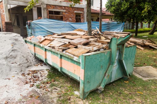 A large, rectangular industrial waste skip situated on a grassy area near a residential street, filled with numerous wooden planks and panels of varying sizes, shapes, and finishes. The skip is made of metal with a weathered green coating, showing signs of dirt and rust, and has reinforced edges and side panels. It is positioned on the pavement adjacent to a pile of fine light-colored rubble or soil. In the background, there is a partially constructed brick building with blue tarps covering parts of the structure and scaffolding, along with a line of trees creating a green canopy. The scene suggests a site for rubbish removal, typical of commercial or construction waste collection, with natural daylight illuminating the area, emphasizing the textures of the wood, metal, and surrounding environment. This image exemplifies waste management activities in Beckenham, where Commercial Waste Removal Beckenham provides rubbish removal services in the local area.